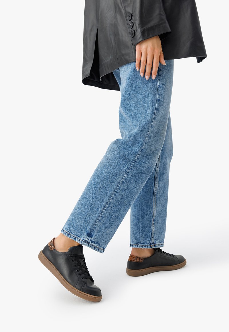 Inimene, kellel on seljas must nahast tossud pruunide aktsentidega, helesinised teksad ja must nahktagi, seistes valge tausta ees.