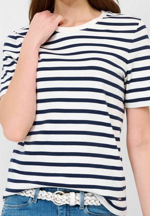 Femme portant un t-shirt à rayures horizontales bleu marine et blanches rentré dans un jean bleu avec une ceinture tressée blanche, main près de la clavicule.