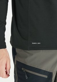 Sorte langærmet top med en tekstureret finish; har "QUICK DRY" label nær kanten. Matchet med flerfarvede bukser, der viser lommehardware.
