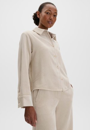Vrouw draagt een beige geribd lang knoopoverhemd en bijpassende broek, handen in de zakken, poseert tegen een effen achtergrond.