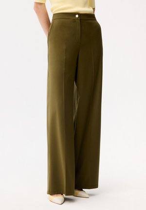 Vrouw draagt een high-waist, olijfgroene wijd uitlopende broek met een crèmekleurige top en beige schoenen met een spits toelopende neus, staand tegen een witte achtergrond.