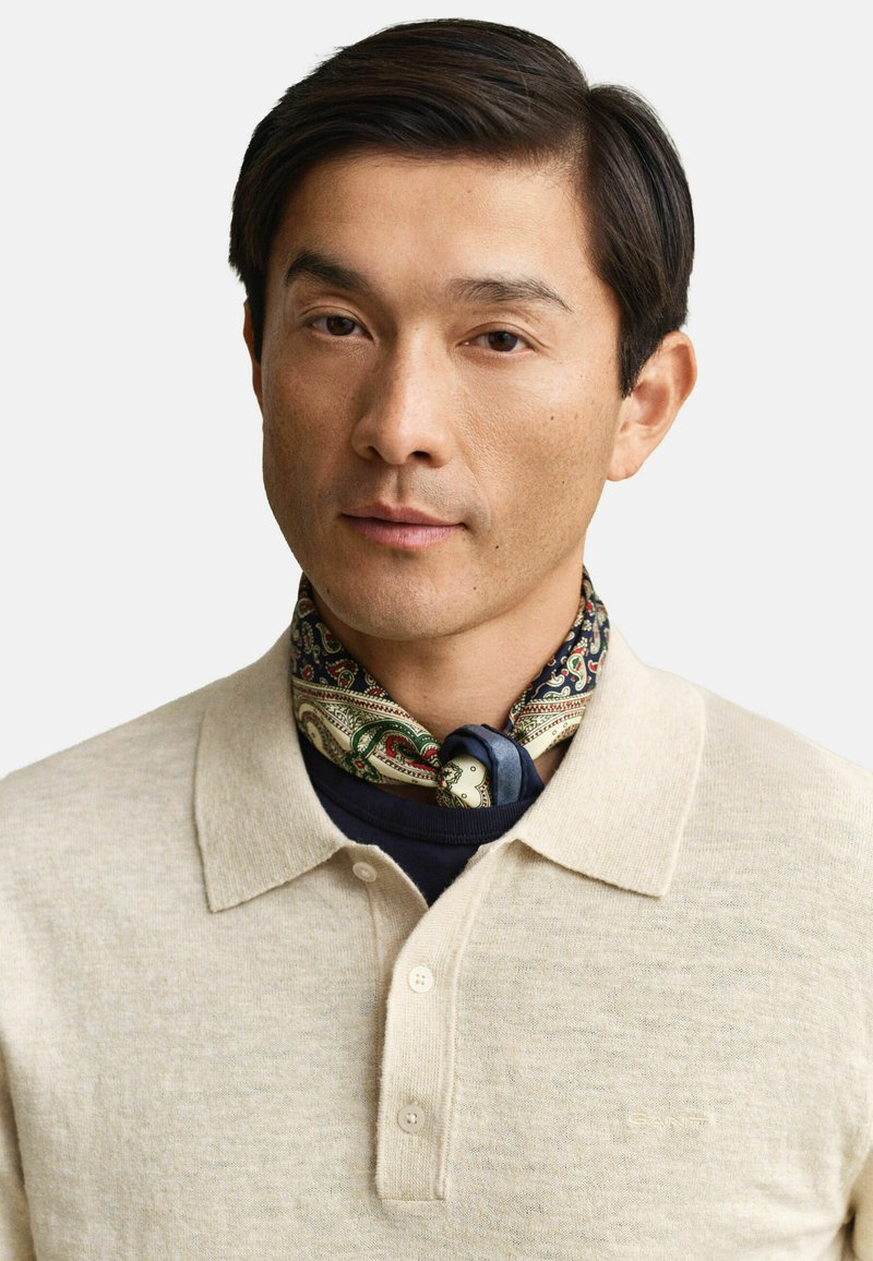 Homme aux cheveux courts et foncés portant un pull beige à col et une écharpe en soie à motifs nouée autour du cou.