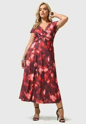 Frau trägt ein rot-schwarz gemustertes Wickelkleid mit kurzen Ärmeln und schwarzen High-Heel-Sandalen, steht mit einer Hand hinter dem Kopf.