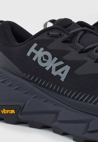 Musta urheilukenkä, jossa on teksturoitu verkko-osa, voimakkaasti pehmustettu sisus, musta kumipohja ja HOKA-logo, jossa on kirkas keltainen Vibram-tunnus.