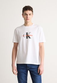 Hvit bomull t-skjorte med en stor svart og oransje "CK" logo med teksten "Calvin Klein Jeans" sentralt foran. Korte ermer, rund hals.