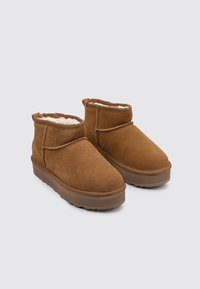 Botas de tobillo de ante marrón con un forro interior suave y esponjoso, que presentan una puntera redondeada y una suela de goma gruesa y texturada.