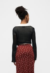 Γυναίκα με σγουρά μαλλιά σε κότσο, φοράει διαφανές μαύρο μπλουζάκι με μακριά μανίκια πάνω από λευκό πουκάμισο και κόκκινη φούστα με λευκά σχέδια καρδιάς, στραμμένη πλάτη.