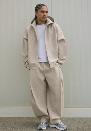 Beige hættetrøje og matchende vide bukser lavet af blødt stof, med en hvid t-shirt nedenunder og grå sneakers med røde detaljer.