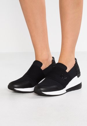 Czarne trampki typu slip-on z siateczkową tkaniną, elastyczny pasek na górze, biała i czarna gumowa podeszwa, noszone na bosych stopach na neutralnym tle.