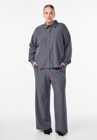 Sieviete stāv ar rokām kabatās, ģērbusies brīvā pelēkā krekliņā ar pogām un atbilstošās platajās biksēs, kājās valkājot melnas lokveida kurpes.