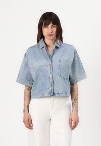 Giacca di jeans blu chiaro corta con maniche corte, bottoni frontali e una tasca sul petto. Texture liscia con una vestibilità comoda.