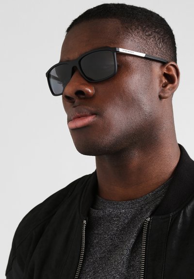 Black sunglasses with rectangular frames, dark lenses, and silver accent on the temple, worn by a person in a black jacket and gray shirt.