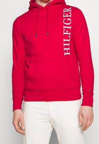 Felpa rossa in cotone con tasca a marsupio. Presenta un cappuccio con coulisse e il testo del logo bianco verticale sul lato sinistro. Polsini e orlo a coste.