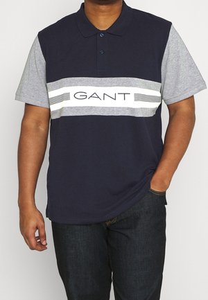 Navy blue polo shirt with gray short sleeves. Features a white horizontal stripe with "GANT" text and lines across the chest. Soft cotton fabric.