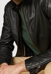 Černá kožená bunda s vysokým límcem, zdobená zlatým zipem a bočními kapsami, nošená přes zelenou košili. Hladká textura a přiléhavý střih.