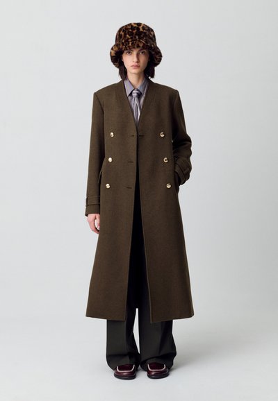 Cappotto in lana verde oliva doppiopetto con bottoni dorati, abbinato a una camicia a righe e un cappello di pelliccia con stampa leopardo marrone.