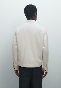 Beige, struktureret jakke lavet af glat stof. Har en klassisk krave og knapdetaljer på ærmerne. Bagside vises.