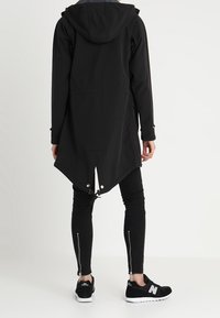 Cappotto lungo nero con cappuccio, dotato di orlo curvo, lacci di regolazione, bottoni a pressione e tasche laterali. Indossato sopra leggins neri e sneakers.