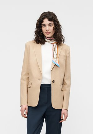 Kvinde med krøllet hår iført en beige blazer, hvid top, marineblå bukser og et farverigt mønstret tørklæde om halsen.