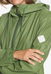 Personne portant une veste de pluie verte avec capuche, fermeture éclair à l'avant, cordons de serrage, et un patch carré blanc avec logo sur la manche, bras croisés.