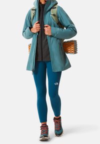 Vattentålig tealjacka med huva, över en svart fleece-topp. Kombinerad med blå leggings och slitstarka, flerfärgade vandringsskor.