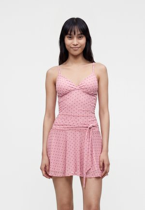 Jeune femme aux longs cheveux foncés portant une robe rose à pois sans manches nouée à la taille, debout devant un fond uni.