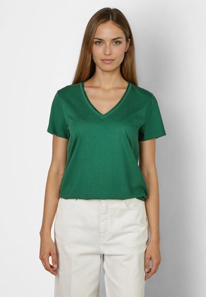 Frau trägt ein grünes V-Ausschnitt-Kurzarmshirt mit kleinem Herzmotiv und weiße Hose, steht vor einem hellgrauen Hintergrund.