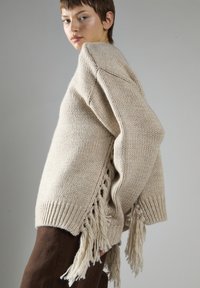 Osoba ubrana w beżowy sweter o grubym splotcie z długimi frędzlami po bokach oraz w brązowe spodnie, na jednolitym szarym tle.