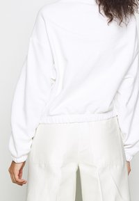 Person som bär en vit långärmad kort sweatshirt och högmidjade vita byxor, sedd från ryggen med lockigt hår synligt.