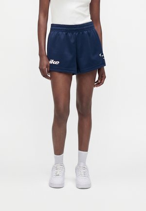 Personne portant un short de sport Nike bleu marine, des baskets blanches et des chaussettes blanches, debout devant un fond clair uni.