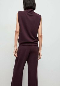 Persona con capelli neri corti che indossa un top senza maniche di un viola scuro e pantaloni larghi abbinati, in piedi contro uno sfondo chiaro e neutro.