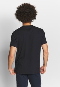 Muž s kudrnatými vlasy, oblečený v jednoduchém černém tričku s krátkým rukávem a námořnických kalhotách s bílými bočními pruhy, otočený zády na bílém pozadí.