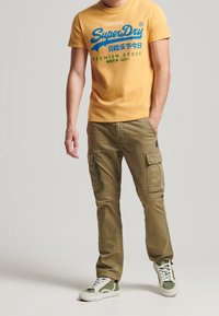 Camiseta amarilla de manga corta con texto gráfico azul y verde, combinada con pantalones cargo oliva con bolsillos laterales y acentos verdes. Zapatillas blancas y verdes.