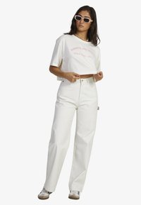 White cropped t-shirt with pink text paired with high-waisted white wide-leg jeans. Model wears white sunglasses and sneakers.