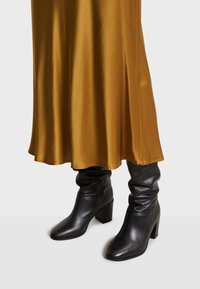 Bottes noires montantes à talons avec une texture lisse et un bout pointu, associées à une robe en satin doré qui tombe avec élégance.