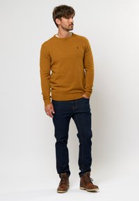 Senfgelber Strickpullover mit Rundhalsausschnitt, gerippten Bündchen und Saum; kombiniert mit dunkelblauen Jeans und braunen Schnürstiefeln.
