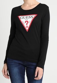 Zwarte lange mouwen shirt met een driehoekige rode en witte afbeelding met "GUESS" en een vraagteken, gemaakt van een zachte stof.