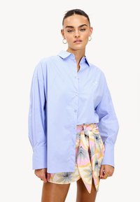 Mujer con el cabello recogido, usando pendientes de aro dorados, una camisa de botones de gran tamaño en azul claro y una falda envolvente floral colorida, de pie contra un fondo blanco.