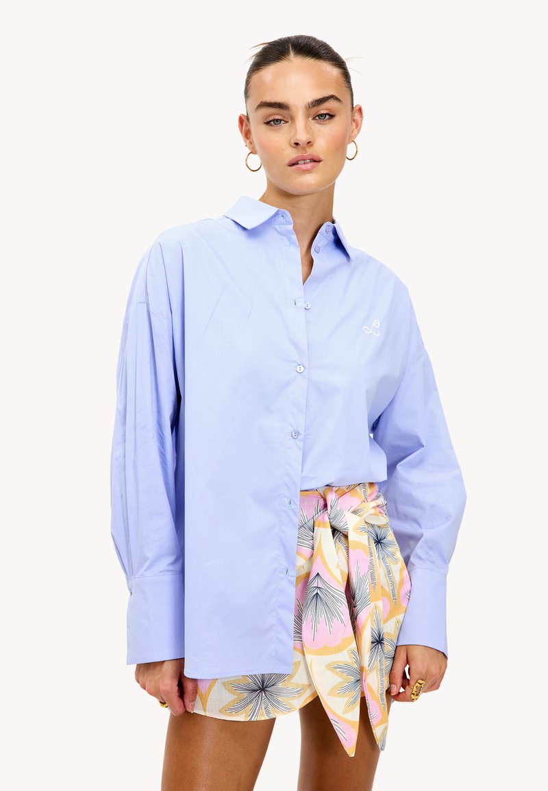 Mujer con el cabello recogido, usando pendientes de aro dorados, una camisa de botones de gran tamaño en azul claro y una falda envolvente floral colorida, de pie contra un fondo blanco.