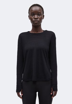 Femme aux cheveux courts portant un haut noir à manches longues et un pantalon noir, debout devant un fond clair uni, regardant vers l'avant.