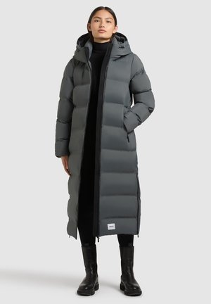 Langer, grauer Puffer-Mantel mit Kapuze, ausgestattet mit einem Frontreißverschluss, gestepptem Design und Seitentaschen. Getragen über einem schwarzen Outfit und Stiefeln.