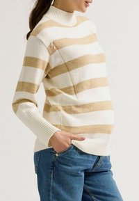 Gestreifter Strickpullover in Creme- und Beigetönen mit hohem Rippkragen. Verfügt über eine Seitentasche und gerippte Bündchen. Getragen mit blauen Jeans.