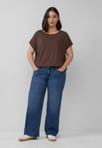 Vrouw die staat met haar handen in de zakken, gekleed in een bruin T-shirt met korte mouwen, wijde blauwe jeans en witte sneakers tegen een effen grijze achtergrond.