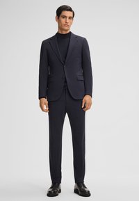 Abito blu navy con un blazer aderente, rever a punta e pantaloni coordinati. Indossato sopra un dolcevita nero. Comprende scarpe nere con lacci.