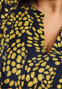 Bluzka z granatowego materiału z dekoltem w kształcie litery V, z żółtym wzorem w owalne plamy i miękką teksturą, z delikatnymi marszczeniami na ramionach.