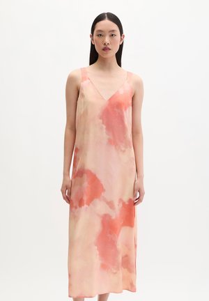 Femme aux longs cheveux noirs portant une robe sans manches à col en V, longueur cheville, avec des motifs aquarelles pêche et corail, debout devant un fond blanc.