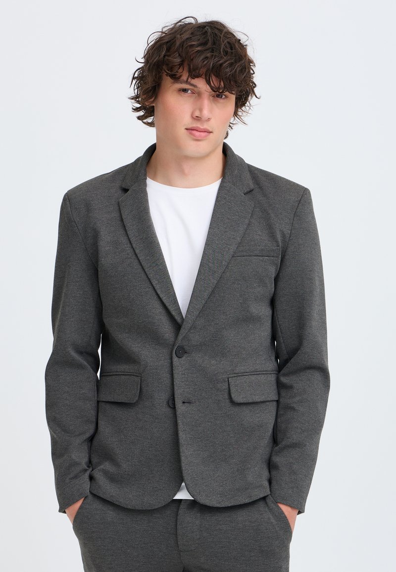 Jeune homme aux cheveux bouclés portant une veste de costume gris foncé et un pantalon assorti sur un t-shirt blanc, mains dans les poches, fond neutre.