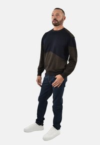 Maglione blu scuro e verde oliva con un motivo geometrico, abbinato a jeans blu scuro e sneaker bianche. Texture lavorata a maglia, scollatura a girocollo.