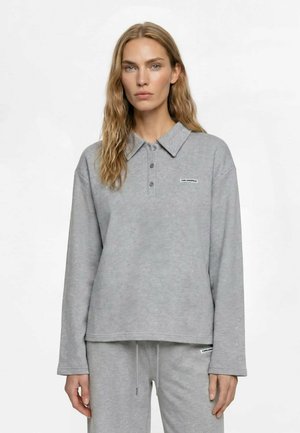 Donna che indossa una polo a maniche lunghe grigio chiaro e pantaloni della tuta abbinati, in piedi su uno sfondo bianco uniforme.