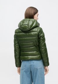 Grøn dunjakke med en skinnende, quiltet tekstur, med hætte og elastisk kant. Kombineret med lyseblå, højtaljede jeans.